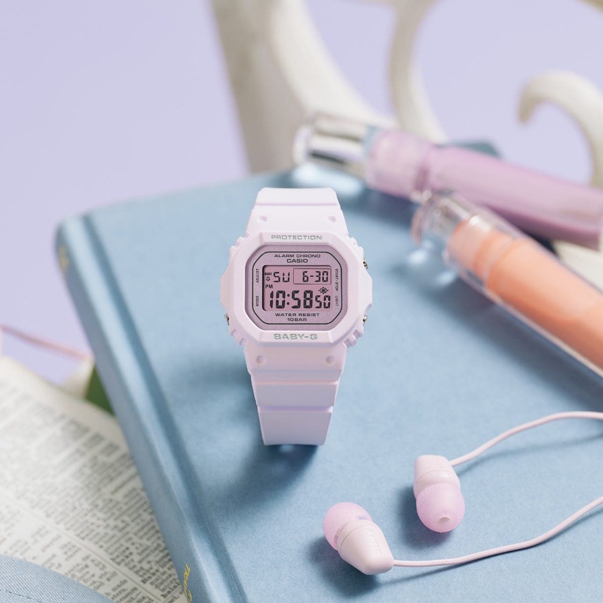 The BABY-G BGD-565SC-4BER digital clock is sitting on a blue book with pens and headphones next to it.
