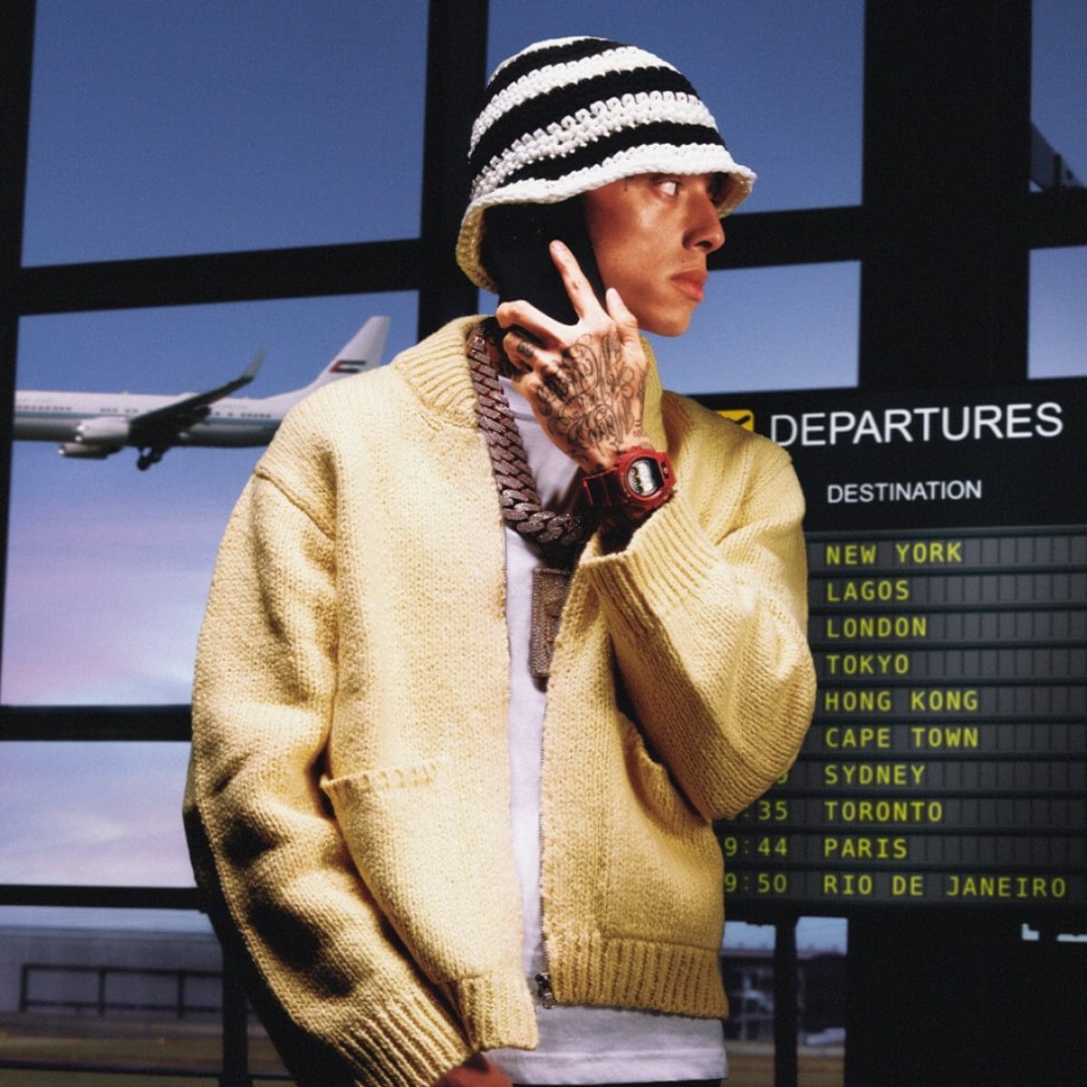 Central Cee stands at an airport gate and talks on the phone. An aeroplane and a display board can be seen in the background. He wears the DW-6900CC25-4 wristwatch from G-SHOCK on his wrist.