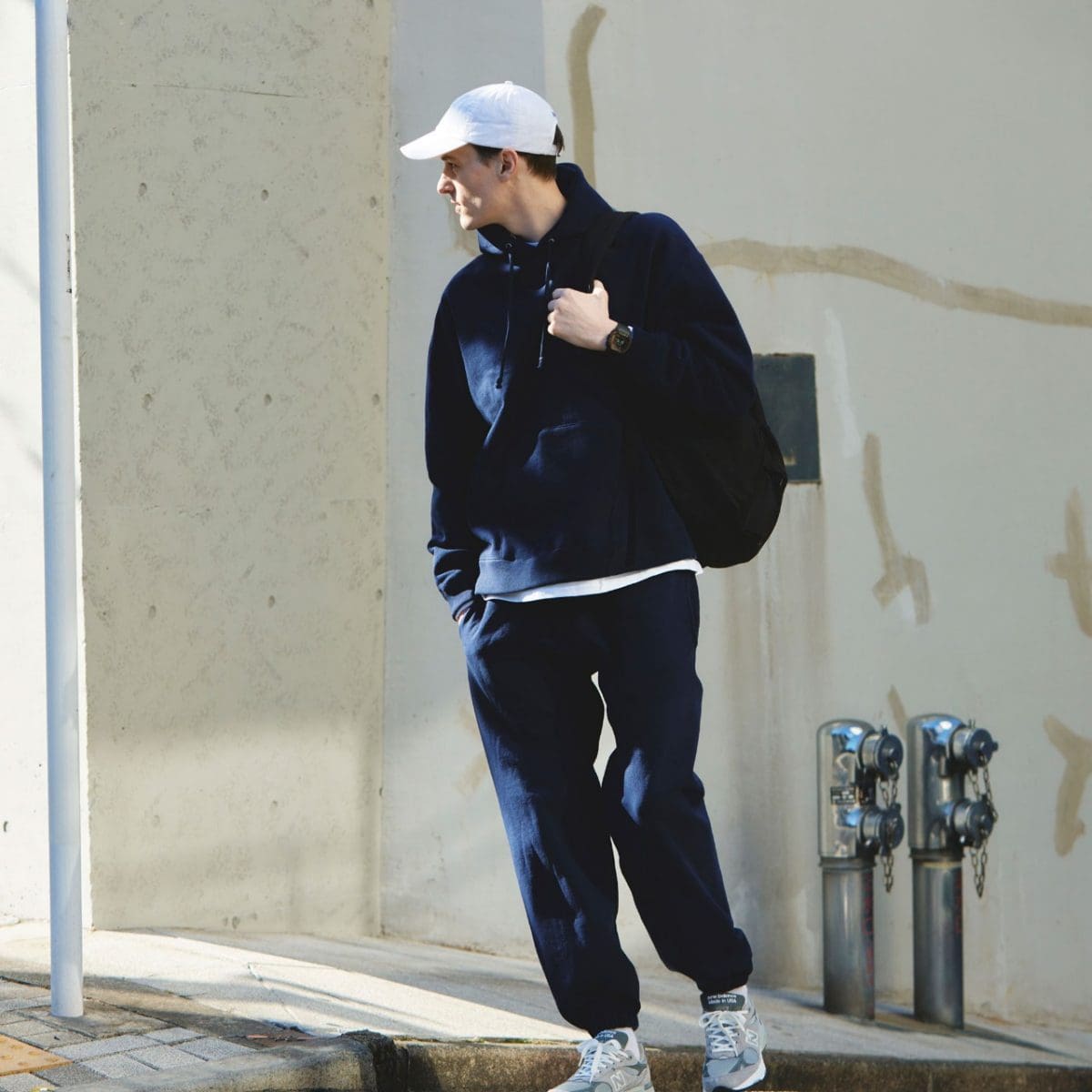 A man in a dark track suit and a white cap is standing at the corner of a building