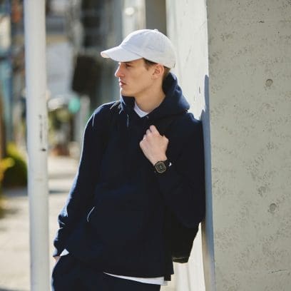 Half-length shot of a man leaning to a building with a dark tracksuit and a white cap, wearing the G-SHOCK DW-H5600 sport watch