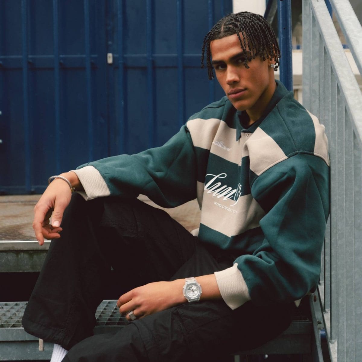 Young man sitting on a metal staircase. He is wearing the GA-2100BM-7A8 in combination with a dark green and beige sweater.