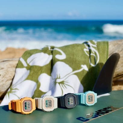 Four surfer digital watches from G-SHOCK: the orange GLX-S5600-4, the GLX-S5600-7B with beige and white strap, the GLX-S5600-1 with grey strap and the GLX-S5600-3 in turquoise on a surfboard on the beach