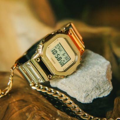 The GM-5600YMG-9 digital watch on a stone next to a gold chain