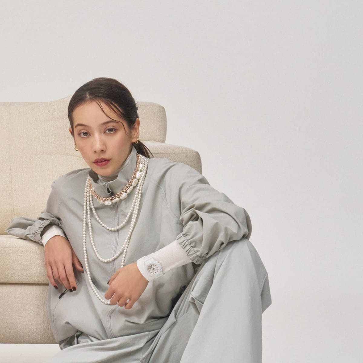 A woman wearing a white sweater and gray jacket leans on a sofa and wears the beige white GMA-P2100M-7A on her wrist