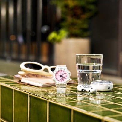 The G-SHOCK GMA-P2100SR-7AER with a transparent strap sits on a green tiled table with summer accessories next to it.