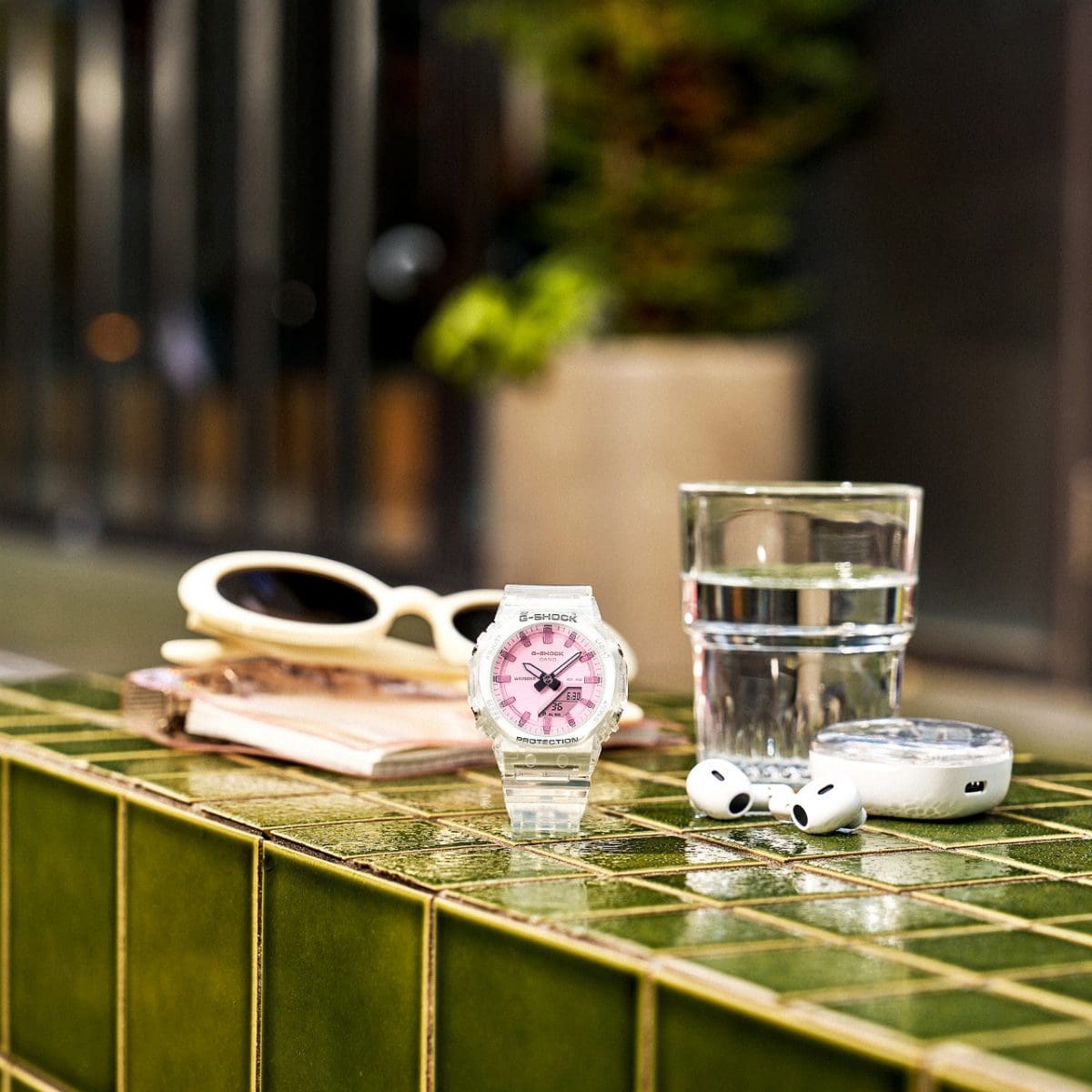 The G-SHOCK GMA-P2100SR-7AER with a transparent strap sits on a green tiled table with summer accessories next to it.