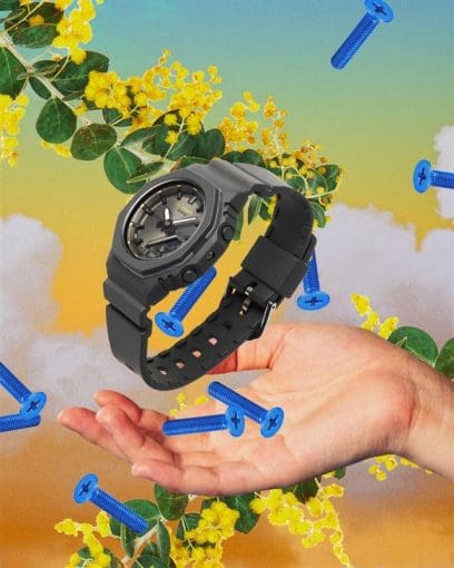 The GMA-P2126W-8AER floats through the air with blue screws and branches with yellow flowers; a hand is held under the watch.