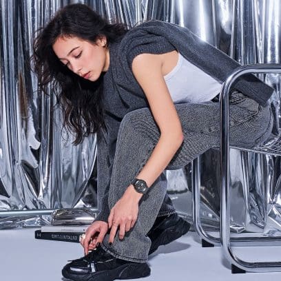 A woman with brown hair and a grey top sits on a metal chair and ties up her black sneakers. On the other wrist she is wearing the black G-SHOCK GM-S2110B-8A.