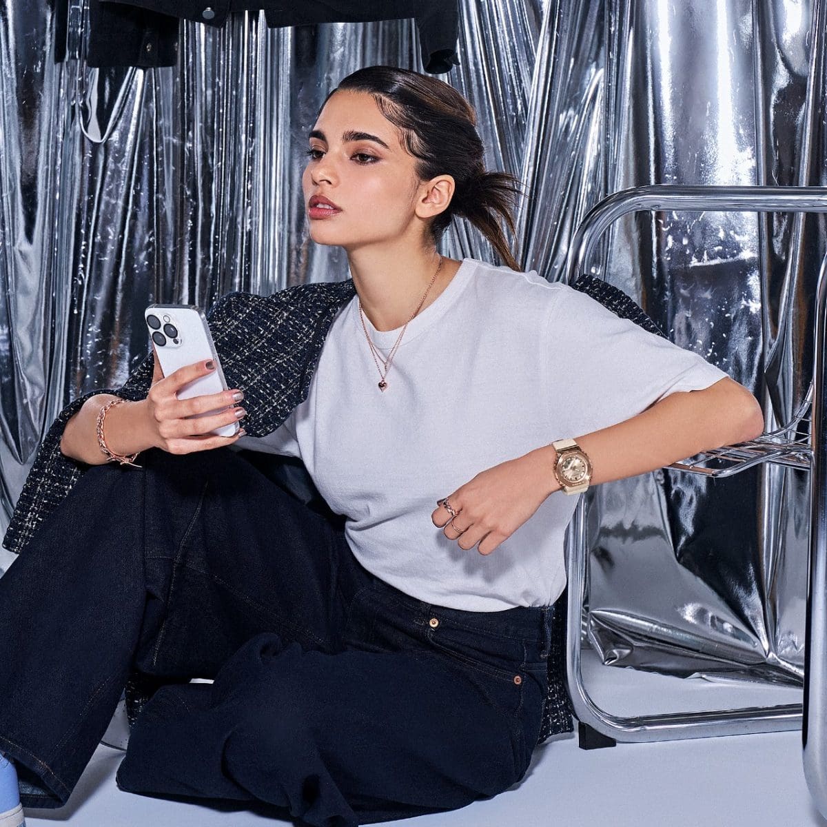 A woman with brown hair and a gray shirt holds a smartphone in her hand and sits on the floor, leaning against a metal chair. On her other wrist she wears the G-SHOCK GM-S2110PG-4A