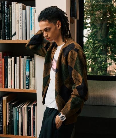 A man wearing a plaid cardigan leans against a bookshelf and wears the GM-2100M-1AER on his wrist.