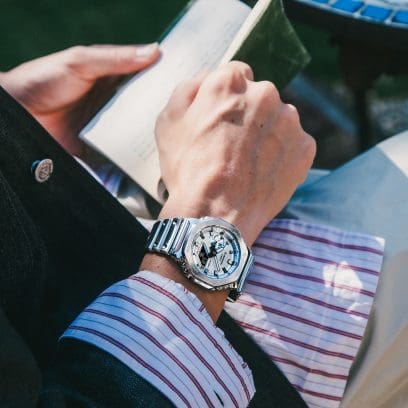 Close-up of the G-SHOCK GM-2100YM-8A wristwatch on the wrist of a man holding a book in his hands.