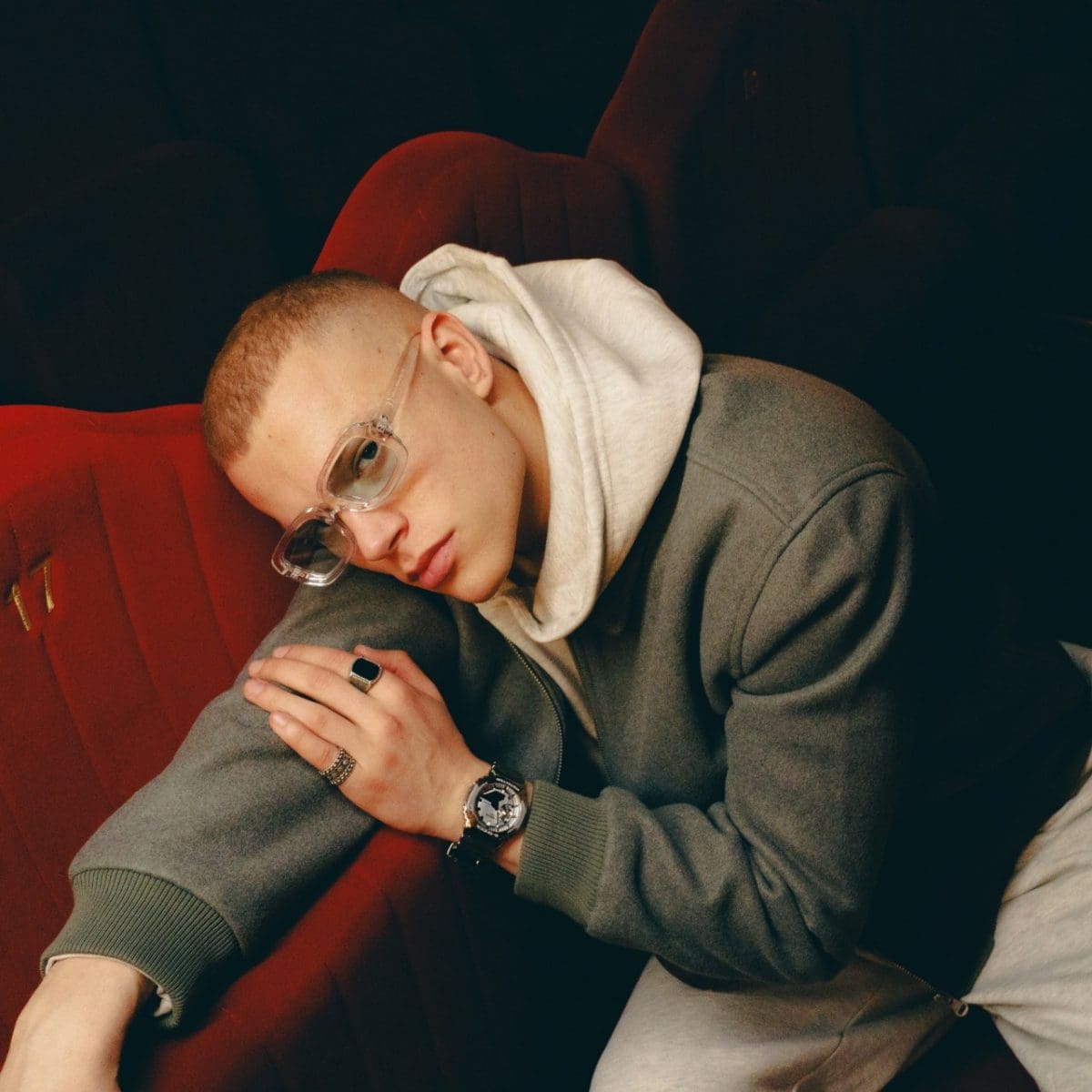 A young man wears the GM-2100YM-8AER on his left wrist, paired with a gray jacket and a white sweater. He sits casually on a red cinema seat and looks into the camera.