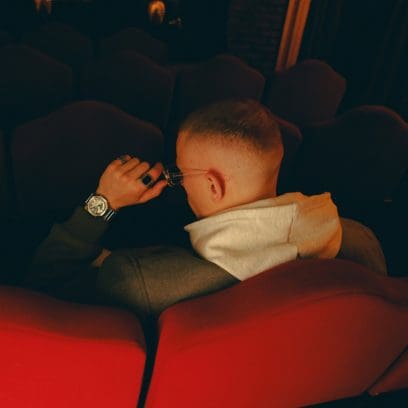 A young man wearing a gray jacket and white sweater sits with his back to the camera on a red cinema seat, reaching for his glasses. He wears the GM-2100YM-8AER on his left wrist, along with two silver rings on his fingers.