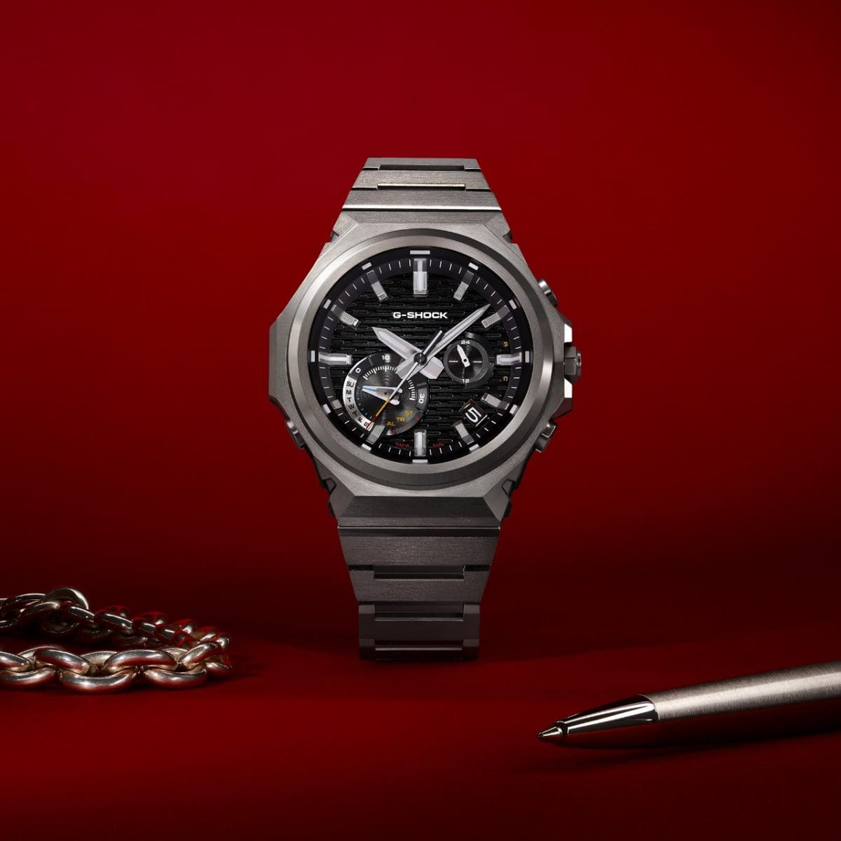 The GST-B1000D-1AER stands against a red background. In front of it lies a silver ballpoint pen and a silver link chain.