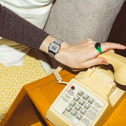 A woman's hand reaches for the handset of an old-fashioned push-button telephone. On her wrist she is wearing the LA680WEL-8 digital watch with a light grey leather strap.