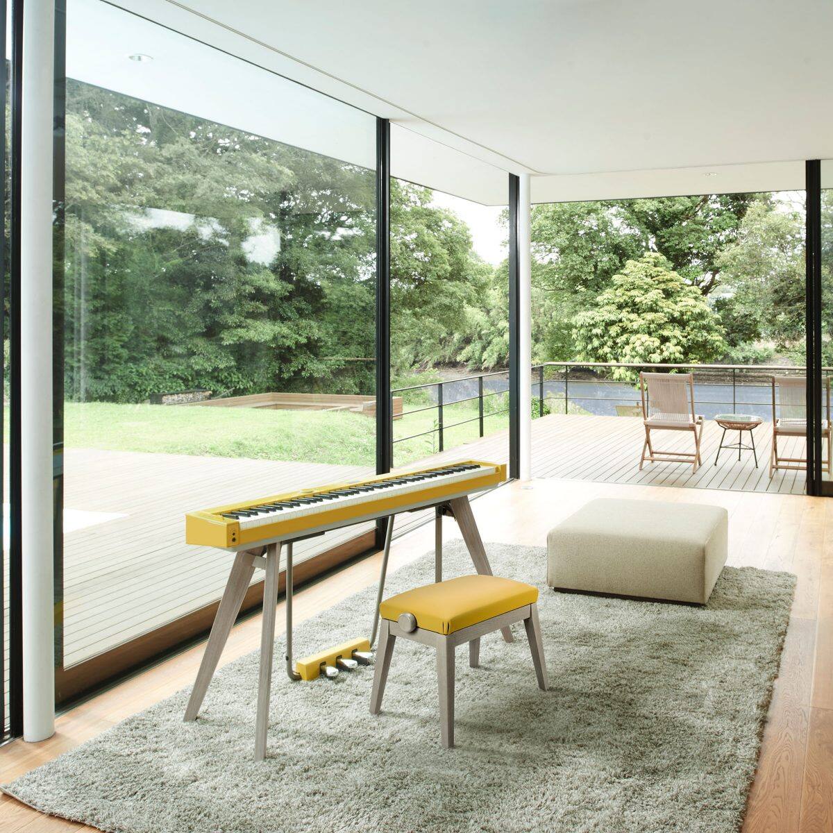 Photo du piano numérique CASIO Privia PX-S7000 avec banquette, coloris Harmonious Mustard, dans un salon au mobilier moderne avec baies vitrées du sol au plafond et vue sur un jardin verdoyant avec terrasse.