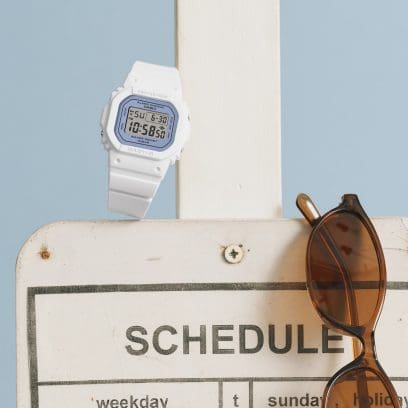 La montre numérique BABY-G BGD-565SC-2BER est appuyée contre un pilier en bois blanc auquel est suspendue une paire de lunettes de soleil.