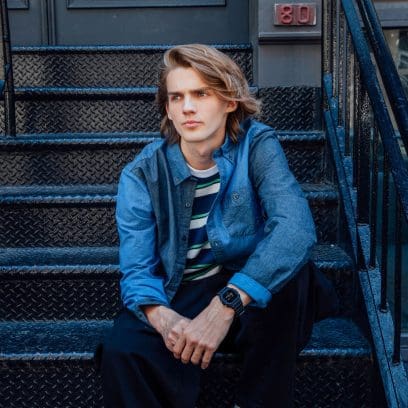 Un homme est assis sur un escalier. Il a les cheveux blonds jusqu'aux épaules, porte une chemise en jean et la montre numérique noire DW-5600 de G-SHOCK.