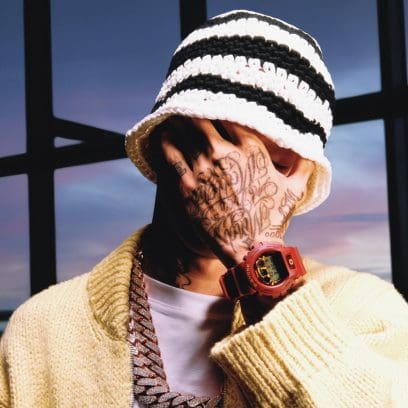 Central Cee cache son visage avec sa main, on aperçoit à son poignet la montre-bracelet DW-6900 rouge foncé.