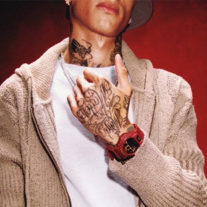 Photo en demi-portrait de Central Cee devant un mur rouge. Il porte une chemise blanche, un cardigan beige et la montre numérique DW-6900 rouge foncé.