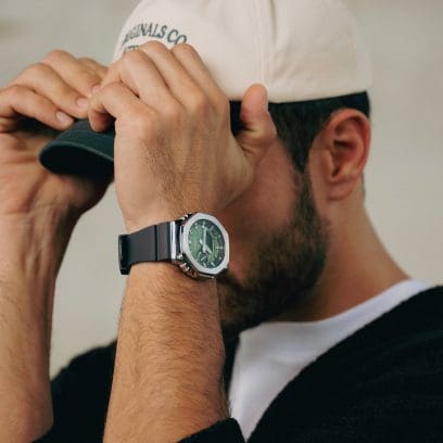 Un homme porte la GBM-2100A-1A3 au poignet, associée à une casquette blanche et verte, un pull noir et un t-shirt blanc. 