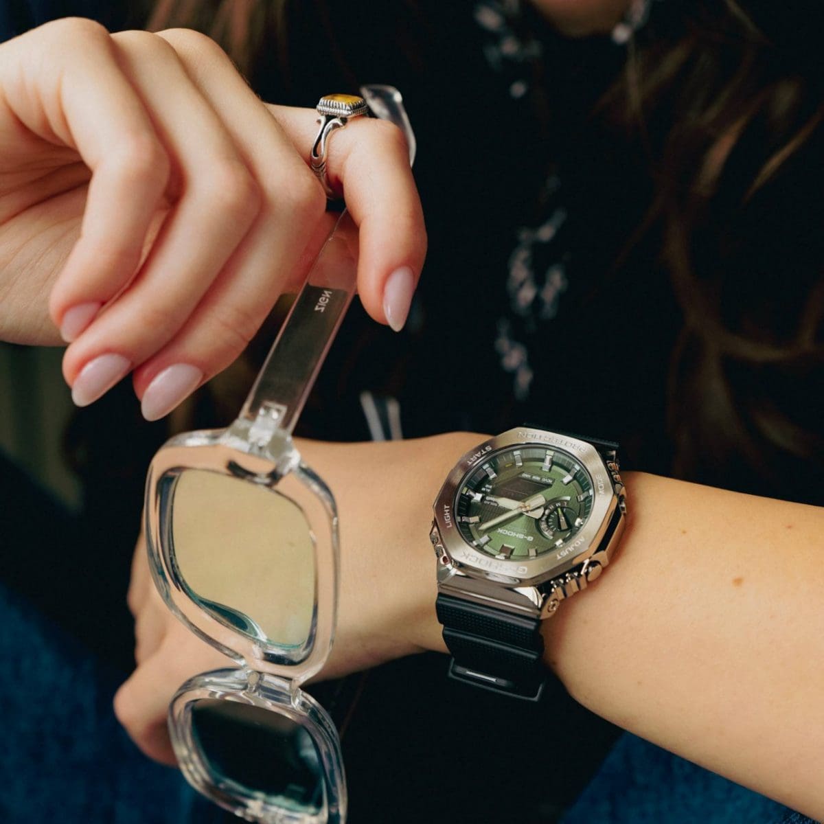 Une femme porte la GBM-2100A-1A3 au poignet, associée à un pull en maille noir et un jean bleu. Elle tient également des lunettes dans sa main droite. 