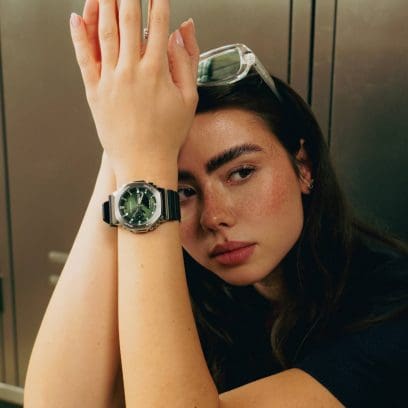 Une jeune femme assise porte la GBM-2100A-1A3 au poignet. Elle porte un pull en tricot noir et a des lunettes sur la tête. 