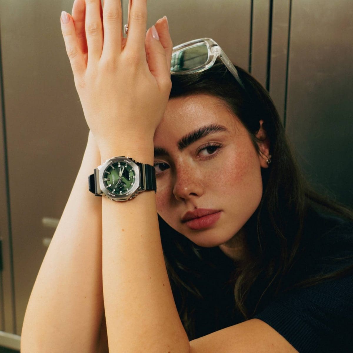 Une jeune femme assise porte la GBM-2100A-1A3 au poignet. Elle porte un pull en tricot noir et a des lunettes sur la tête. 