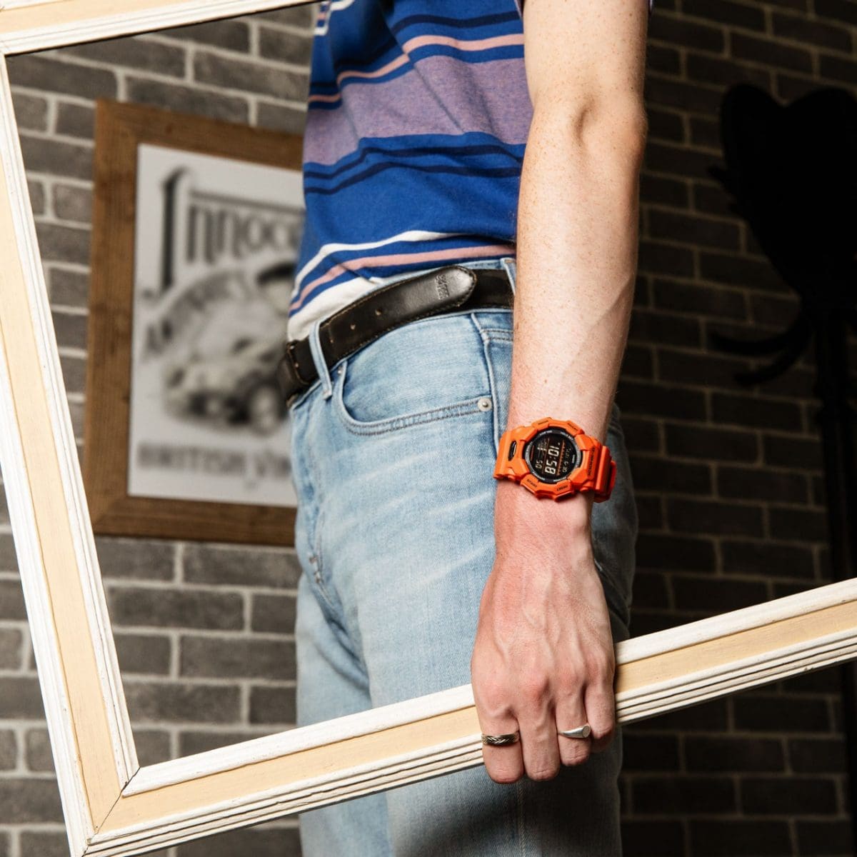 Demi-plan latéral d'un homme portant un jean, un t-shirt à rayures multicolores et la montre-bracelet G-SHOCK GD-010-4 orange.