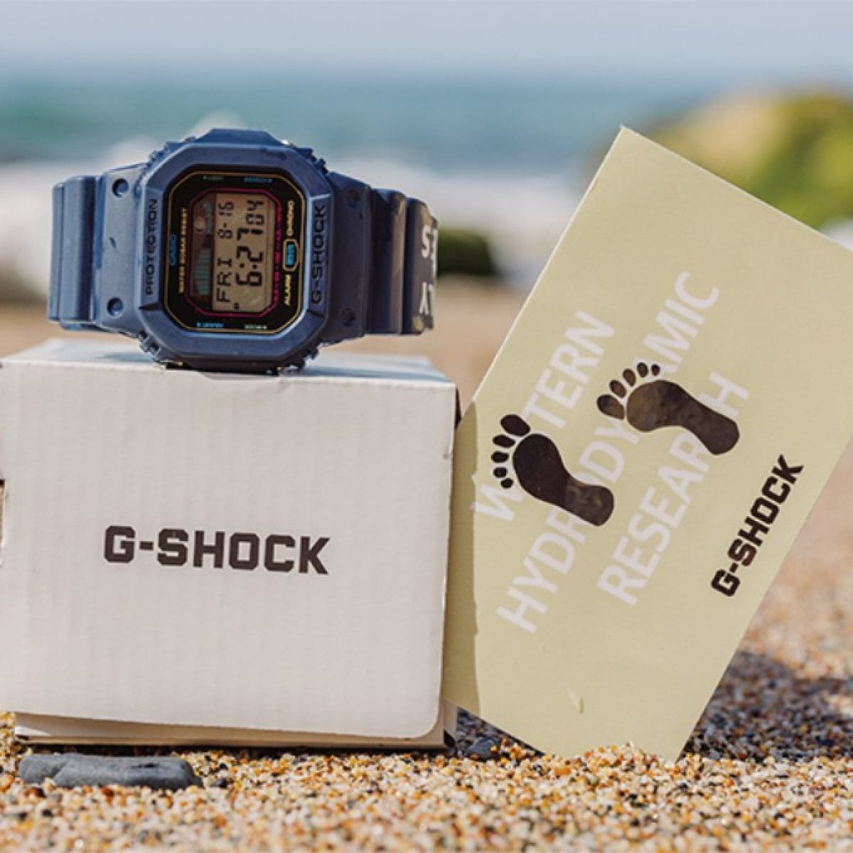 La montre de collaboration G-SHOCK G-LIDE déposée dans le sable sur son emballage en carton.
