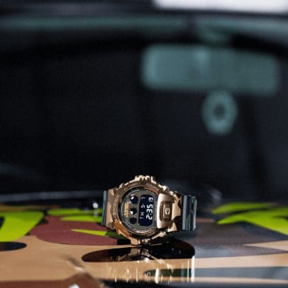 La montre-bracelet GM-6900CMG-3ER, dotée d'un boîtier doré, repose sur un fond au motif camouflage dans des tons bruns et jaunes