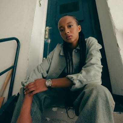 Une jeune femme est assise sur un escalier et porte la GM-S2110-4AER au poignet gauche. Elle associe son look à une veste et un pantalon en jean vert-gris.