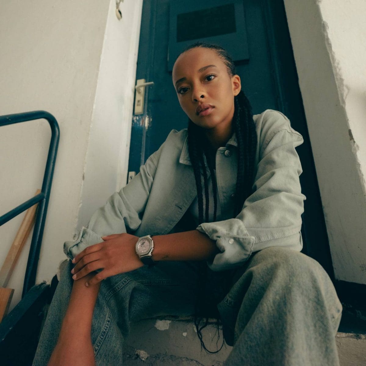 Une jeune femme est assise sur un escalier et porte la GM-S2110-4AER au poignet gauche. Elle associe son look à une veste et un pantalon en jean vert-gris.