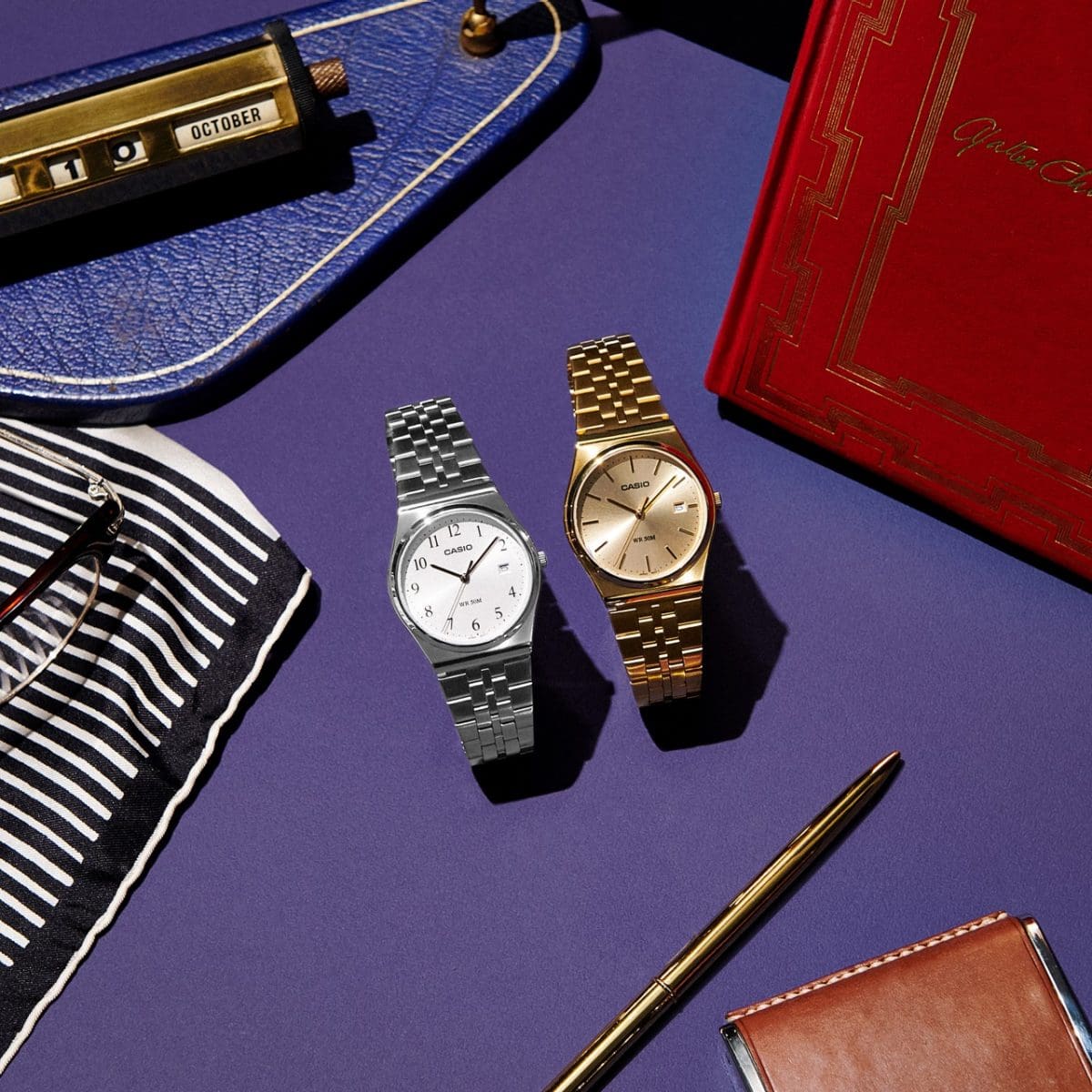 La MTP-B145D-7BV avec bracelet argenté sur un bureau à côté de la MTP-B145G-9AV dorée, d'un stylo, d'un porte-monnaie et d'une trousse rouge.