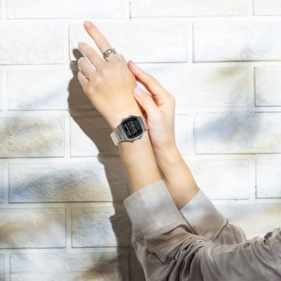 Primo piano dei polsi di una donna che indossa l'orologio digitale A168XESW-9A e posa davanti a un muro di una casa bianca.