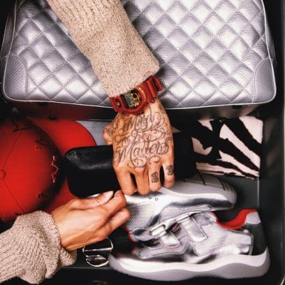 Vista dall'alto del contenuto di una valigia, accanto a un beauty case argentato ci sono delle sneakers argentate, Central Cee prende un astuccio di pelle nera, al polso indossa l'orologio rosso della collaborazione.