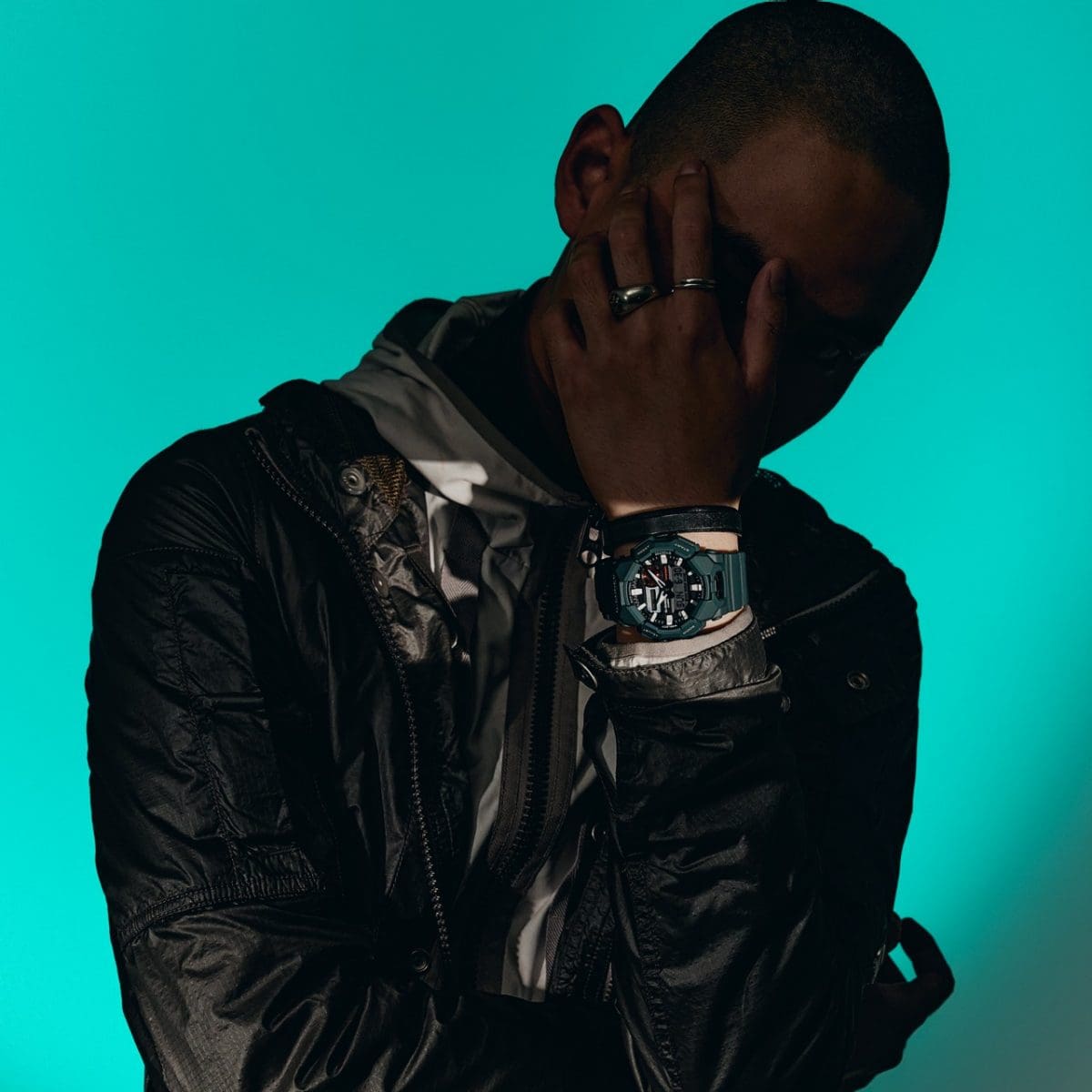 Un uomo si trova davanti a uno sfondo turchese, indossa abiti neri, anelli neri e al polso l'orologio da polso analogico-digitale GA-010-2A di G-SHOCK, di colore verde scuro.