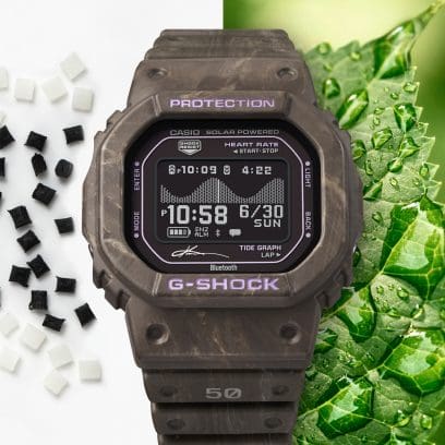 Accanto all'orologio da polso GBX-H5600KI-5ER si vede una foglia verde con gocce d'acqua e piccole palline della materia prima