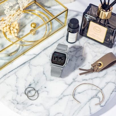 A1000M-1B digitaal horloge met mesh roestvrijstalen band op marmeren blad omringd door sieraden, huisparfums en accessoires