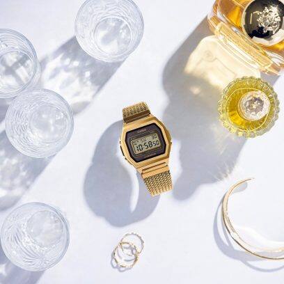 Bovenaanzicht van het goudkleurige roestvrijstalen horloge A1000MGA-5, dat met gesloten band op een witte tafel ligt. Ernaast staan glazen en een fles met een amberkleurige vloeistof. 