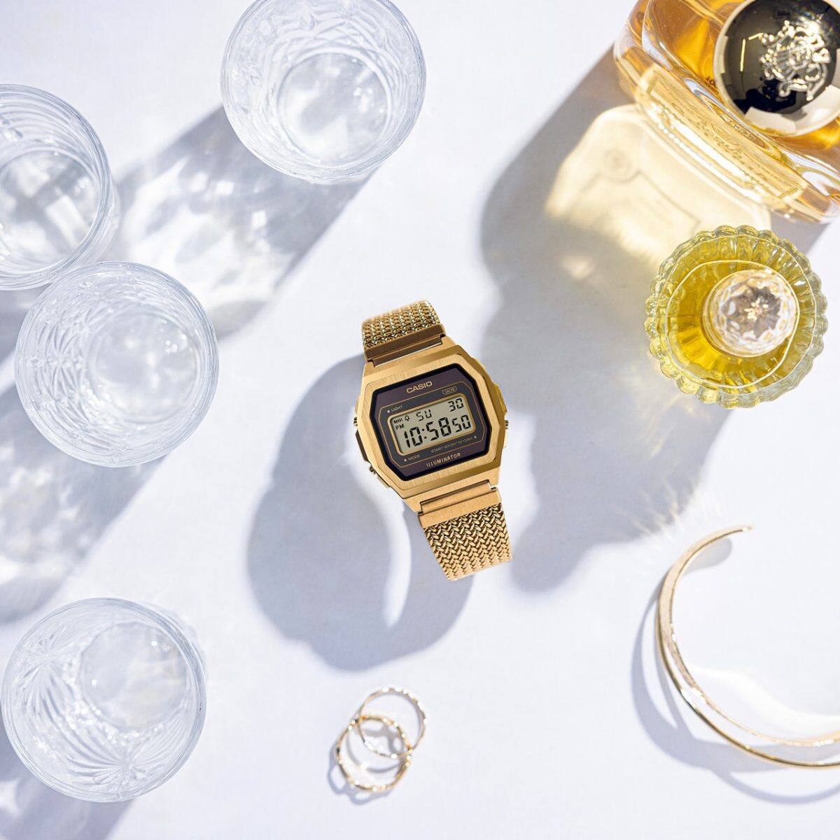 Bovenaanzicht van het goudkleurige roestvrijstalen horloge A1000MGA-5, dat met gesloten band op een witte tafel ligt. Ernaast staan glazen en een fles met een amberkleurige vloeistof. 
