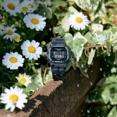 De BGD-565U-1ER op een roestige reling, omringd door witte bloemen, dicht groen gebladerte en vallende waterdruppels.
