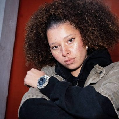 Een vrouw met een zwarte hoodie zit voor een rode muur en draagt ​​de GA-2100CM-5AER om haar pols.