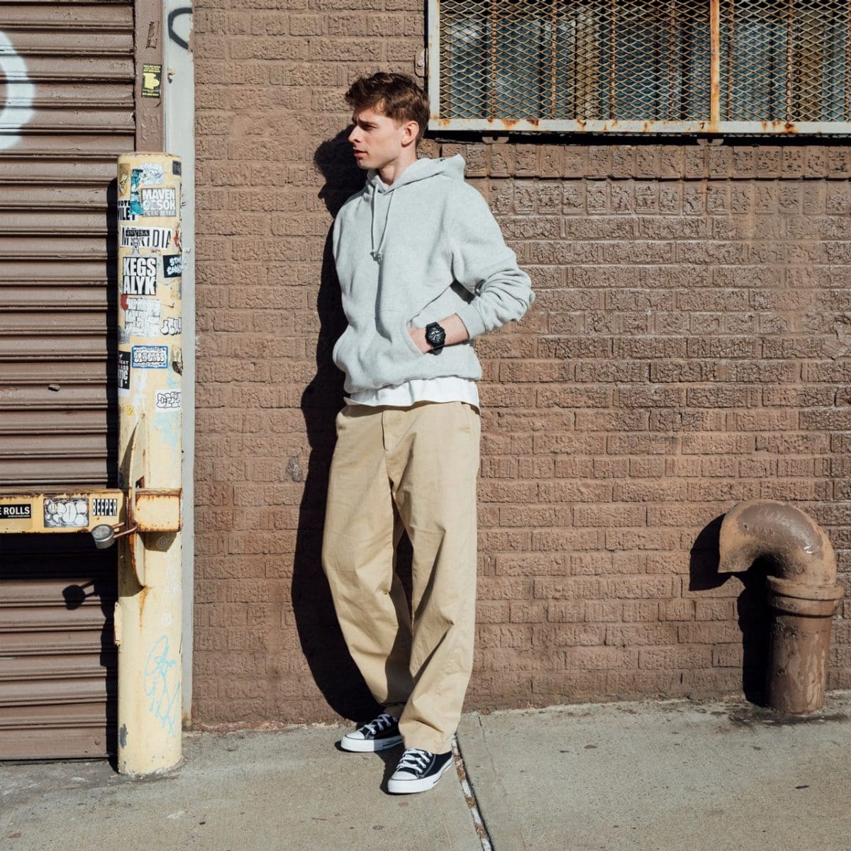 Een man staat in de zon, leunend tegen de muur van een huis en draagt ​​een grijze hoodie, een beige broek en zwarte sneakers. Om de pols is het GA-010-1A1 horloge van G-SHOCK te zien.