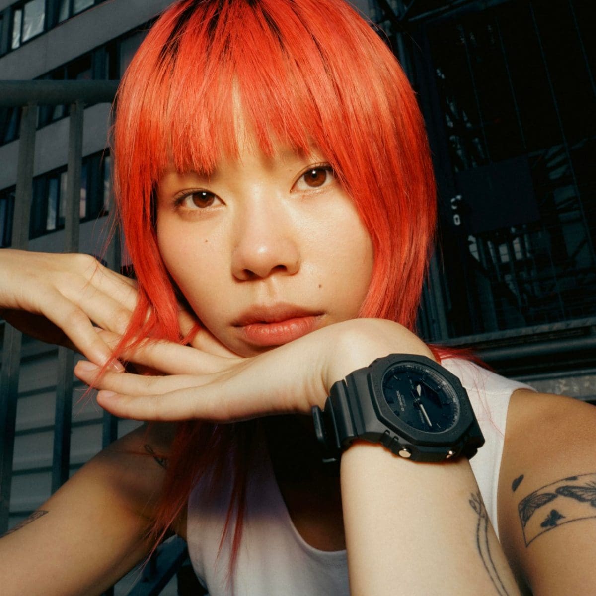 Een vrouw met rood haar kijkt recht in de camera en rust met haar hoofd op haar hand, terwijl ze de GMA-P2100BB-1A om haar pols draagt.