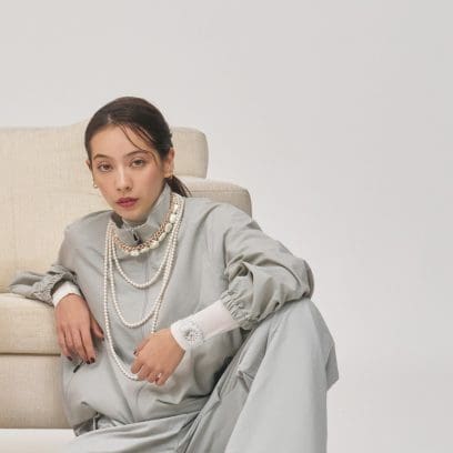 Een vrouw, gekleed in een witte trui en een grijs jasje, leunt op een bank en draagt ​​de beige-witte GMA-P2100M-7A om haar pols