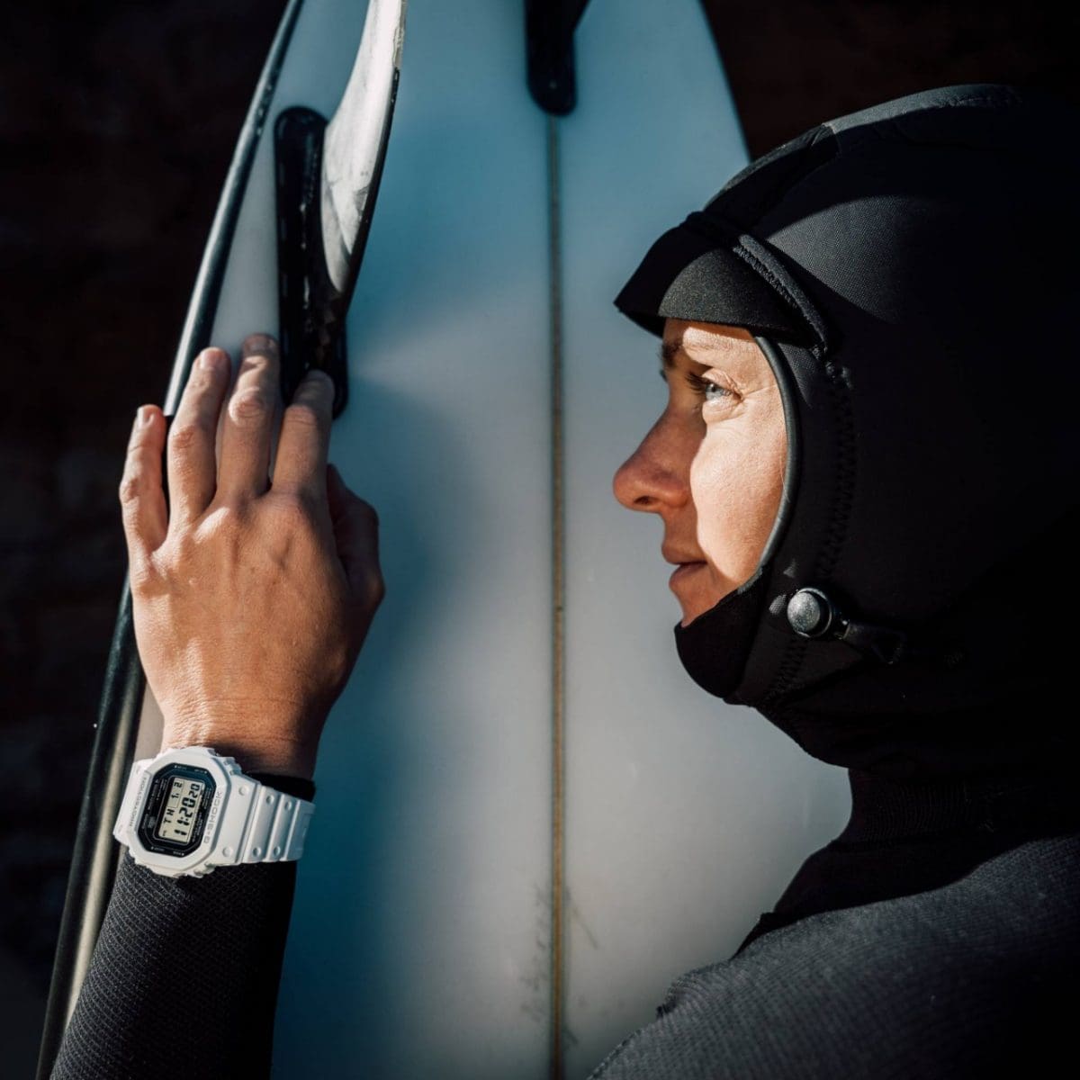 Een vrouw in een wetsuit staat naast een surfplank en draagt ​​de witte GW-5000HS-7ER met een zwarte wijzerplaat om haar pols