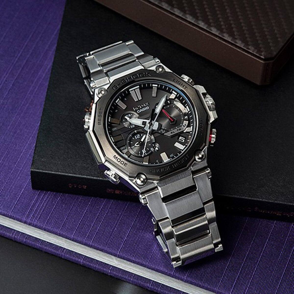 Het CASIO MTG-B2000D-1A polshorloge met zwarte wijzerplaat, zilverkleurige roestvrij stalen kast en zilverkleurige roestvrij stalen armband rust op verschillende notebooks in paars en zwart