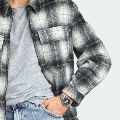 O homem está vestindo uma camisa xadrez preta e branca e uma camiseta cinza. No pulso ele usa um relógio redondo de prata A171WE-1A com pulseira de aço inoxidável