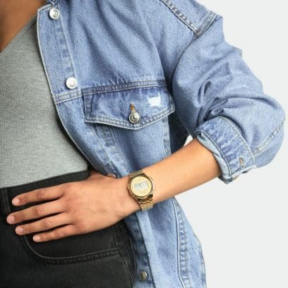 A mulher usa um relógio de pulso redondo de malha dourada combinado com uma jaqueta jeans e um top cinza